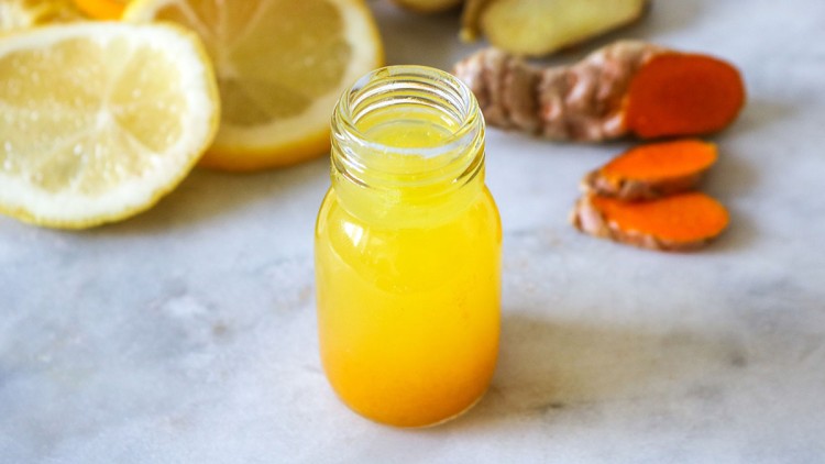 Kleines Glas mit gelbem Saft, im Hintergrund aufgeschnittene Zitronenscheiben, Ingwer und Kurkumawurzel auf heller Oberfläche.