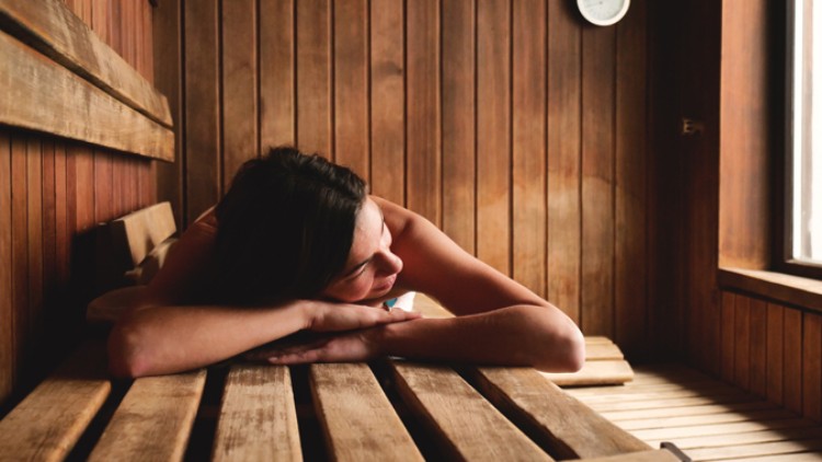 Person liegt entspannt auf einer hölzernen Sitzbank in einer Sauna mit Holzwänden und einem kleinen Fensterausschnitt.