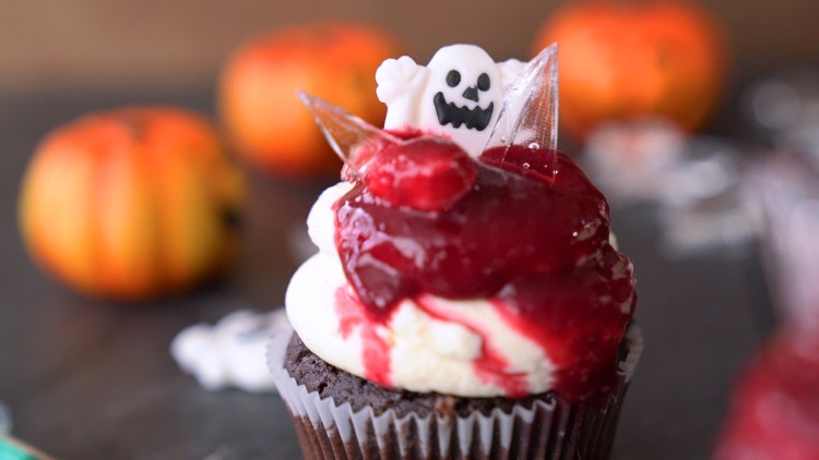 Ein Schokoladen-Cupcake mit weißer Creme, roter Glasur und Dekoration in Form eines weißen Geists vor unscharfen Kürbissen.