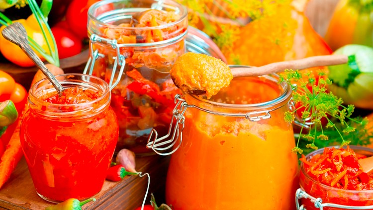 Verschiedene Einmachgläser mit roter und orangefarbener Sauce stehen dekorativ zwischen frischem Gemüse und Kräutern.