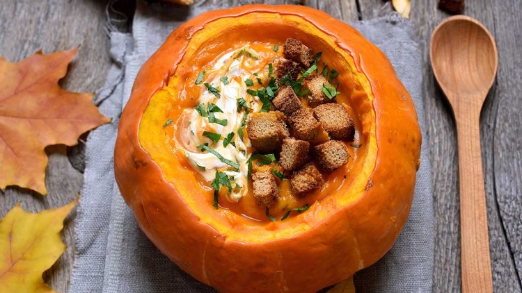 Eine ausgehöhlte, orangefarbene Kürbishälfte mit Suppe, Croutons, Sahne und Kräutern, serviert auf einem grauen Stofftuch.