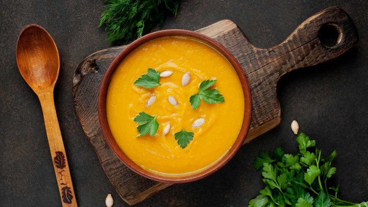Eine Schale mit orangefarbener Suppe, garniert mit Kürbiskernen und Petersilie, auf einem Holzbrett neben Kräutern und einem Holzlöffel.