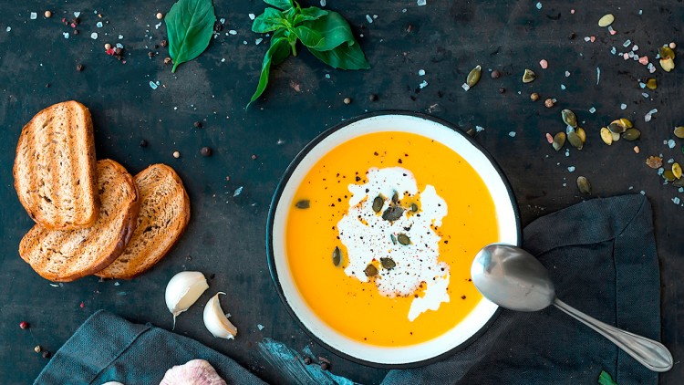 Schale mit oranger Kürbissuppe, Kürbiskernen und Sahne, daneben geröstete Brotscheiben, Basilikum und Knoblauch auf dunklem Untergrund.