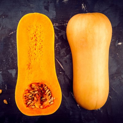 Links ein halbierter Butternut-Kürbis mit sichtbaren Kernen, rechts ein ganzer Butternut-Kürbis auf dunklem Untergrund.