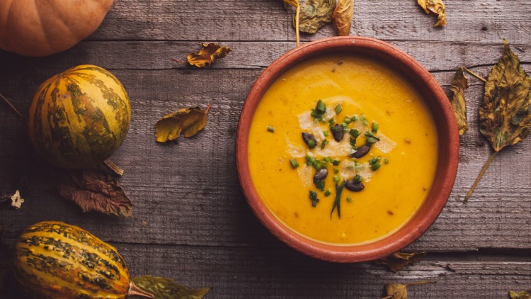 Eine orange Kürbissuppe in einer braunen Schale mit Kürbiskernen und Kräutern, umgeben von Kürbissen und Herbstblättern auf Holztisch.