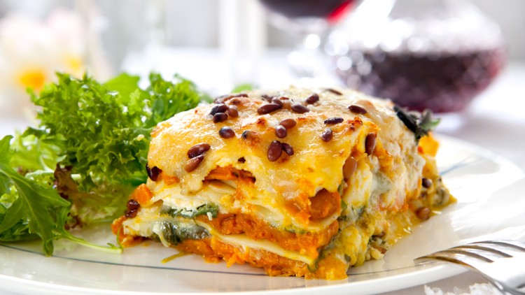 Ein Stück Lasagne mit Käsekruste und Kürbiskernen, serviert auf einem Teller mit frischem Salat, im Hintergrund Rotwein.