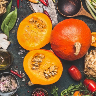 Ein aufgeschnittener orangefarbener Kürbis liegt mit Kernen umgeben von frischen Zutaten wie Chili, Tomaten und Kräutern.