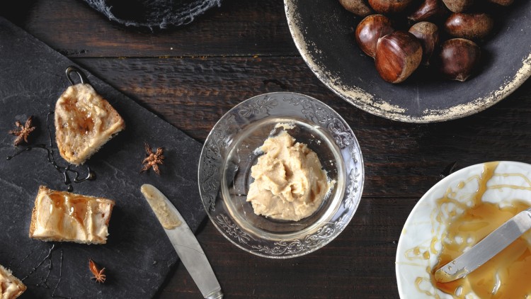 Auf einem Holzuntergrund stehen Maronen, ein Glas mit Creme, Honigbrotstücke, Sternanis und ein Messer mit Creme.