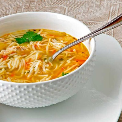 Weiße Schale mit Nudelsuppe, Löffel darin, daneben ein Stück Brot, alles auf einem weißen Teller und beigem Tischtuch.