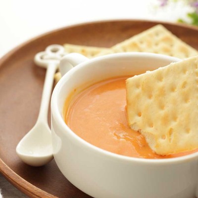Eine weiße Suppenschale mit orangefarbener Suppe, in die ein salziger Cracker getaucht wird, auf einem Holztablett mit Löffel.