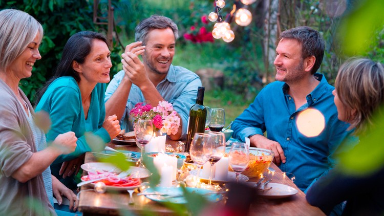 Eine festlich gedeckte Tafel im Garten mit Kerzen, Blumen, Gläsern, einer Weinflasche und Speisen, umgeben von Pflanzen.