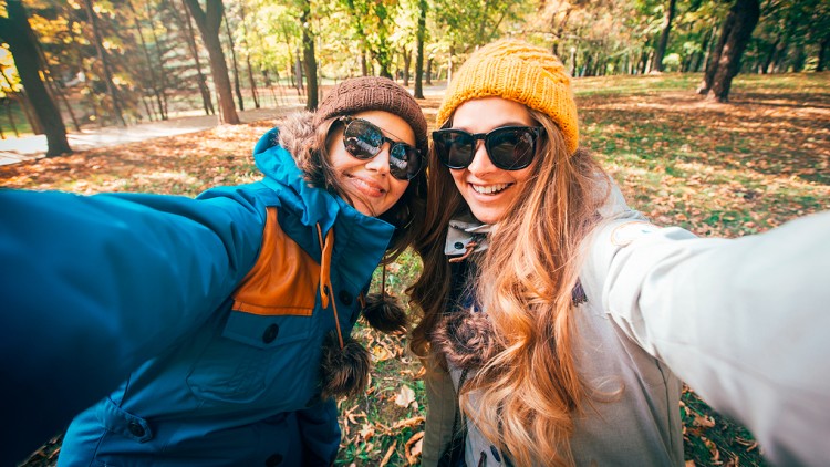 Zwei Personen in Herbstkleidung stehen in einem Park mit Laub am Boden und machen ein Selfie, Bäume im Hintergrund.