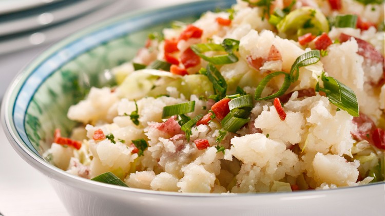 Kartoffelsalat in einer weißen Schüssel mit grünen Zwiebelringen, roten Paprikastückchen und Kräutern, im Hintergrund ein Tellerstapel.
