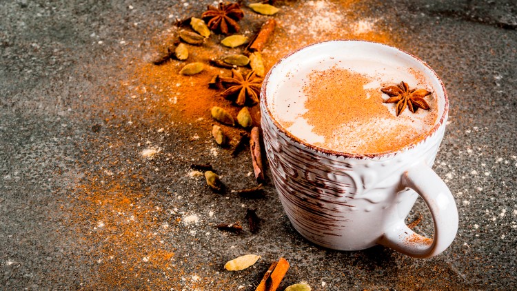 Eine weiße Keramiktasse mit Milchschaum, bestreut mit Zimt, daneben Sternanis, Kardamom und Zimtstangen auf grauem Untergrund.