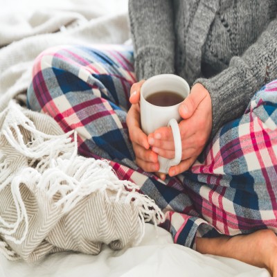 Jemand sitzt im Schneidersitz auf einer Decke mit karierter Hose, hält eine weiße Tasse mit Tee und trägt einen grauen Strickpullover.