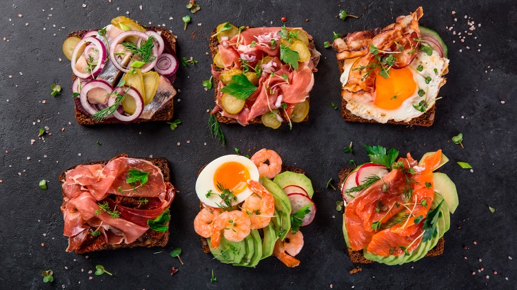 Sechs belegte Brotscheiben mit verschiedenen Toppings wie Fisch, Schinken, Ei, Gemüse und Garnelen auf dunklem Untergrund.