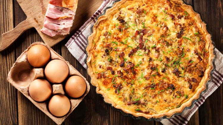 Eine goldbraun gebackene Quiche in einer runden Glasform neben einem Eierkarton mit sechs braunen Eiern auf Holzuntergrund.