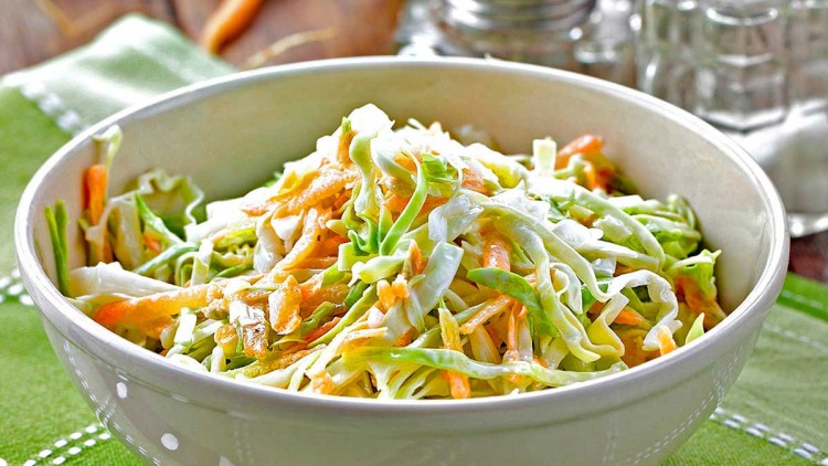 Schüssel mit Krautsalat und Karottenstreifen auf grünem Tuch, im Hintergrund Kohl, Karotten und Salz- sowie Pfefferstreuer.