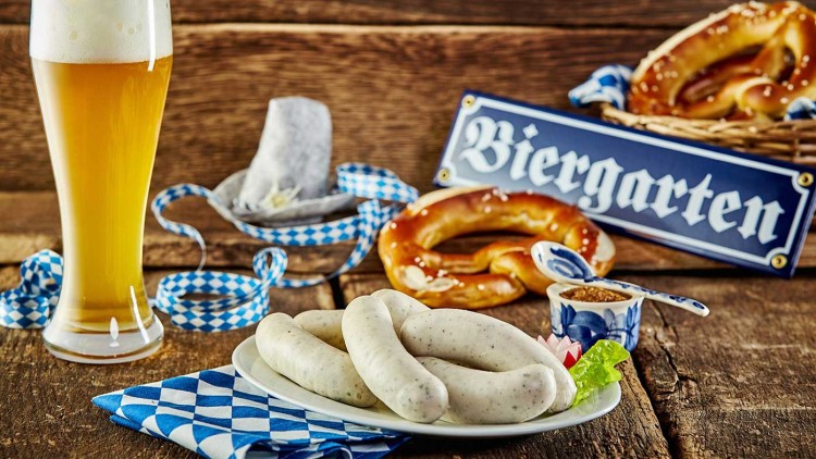 Weißbier, Weißwürste auf einem Teller mit Brezel und Senf, blau-weißes Dekor, Schild mit der Aufschrift Biergarten im Hintergrund.