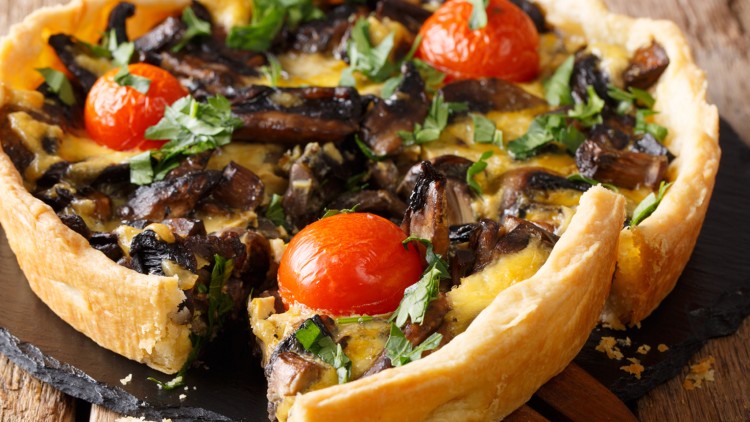 Herzhafte Quiche mit braunen Champignons, Kirschtomaten, gehackter Petersilie und knusprigem Teigrand auf einem Holzbrett.