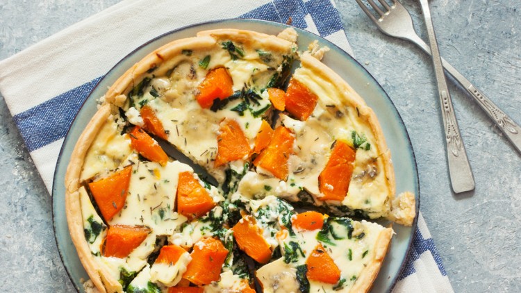 Quiche mit Kürbisstücken, Spinat und Kräutern auf einem grauen Teller, daneben eine gestreifte Serviette und zwei Gabeln.