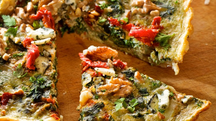 Stück einer Quiche mit Spinat, Walnüssen, getrockneten Tomaten, Petersilie und geschmolzenem Käse auf einem Holzbrett.