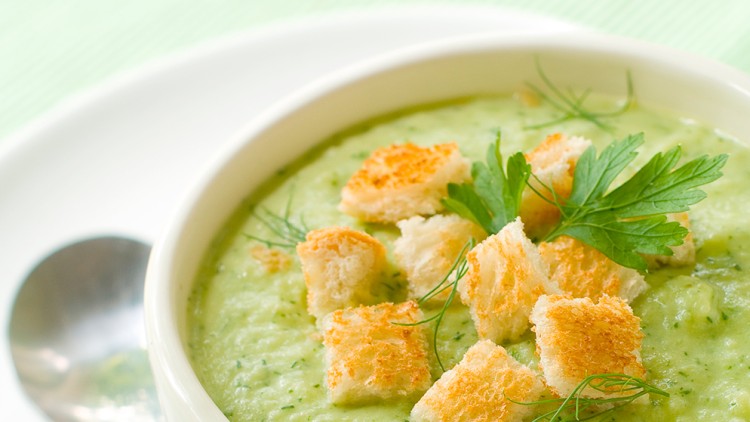 Weiße Schale mit grüner Cremesuppe, belegt mit goldbraunen Croutons, Dill und Petersilie, daneben ein Löffel auf weißem Unterteller.