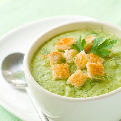 Weiße Schale mit grüner Cremesuppe, belegt mit goldbraunen Croutons, Dill und Petersilie, daneben ein Löffel auf weißem Unterteller.