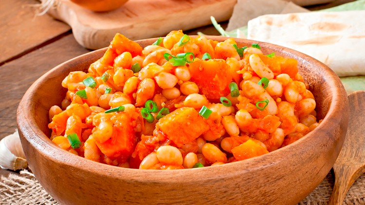 Holzschale mit einem Eintopf aus weißen Bohnen, Kürbis und grünen Kräutern, im Hintergrund Zwiebeln, Tomaten und Fladenbrot.
