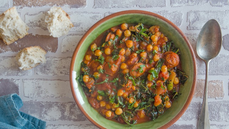 Grüne Schale mit Kichererbsen, Tomatensoße, Gemüse und Kräutern, daneben Löffel, Brotstücke und gefaltetes Tuch.