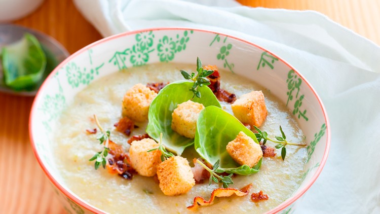 Zwei Schalen mit cremiger Suppe, garniert mit Croutons, grünen Blättern und frischen Kräutern, auf einem Holztisch.