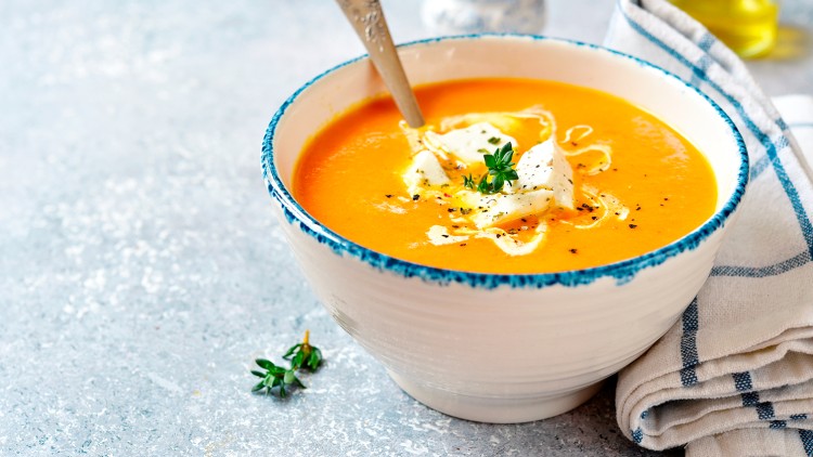 Eine orange Suppe in einer Schale mit blauem Rand, garniert mit Sahne, Kräutern und Käsewürfeln, daneben ein Löffel und Stofftuch.