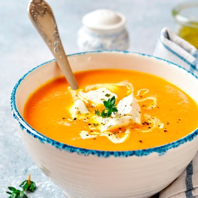 Eine orange Suppe in einer Schale mit blauem Rand, garniert mit Sahne, Kräutern und Käsewürfeln, daneben ein Löffel und Stofftuch.