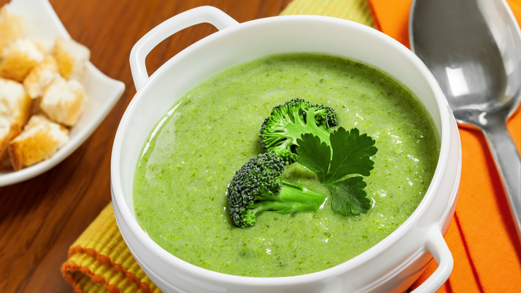 Weiße Schüssel mit grüner Brokkolicremesuppe, garniert mit Brokkoliröschen und Petersilie, neben Croutons und einem Löffel.