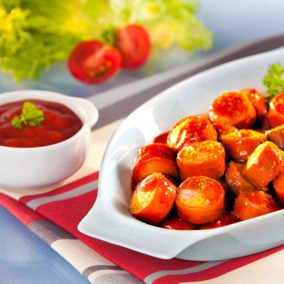 Eine weiße Schale mit Currywurststücken, daneben Pommes, Mayo, Ketchup, Salat und Tomaten auf einem rot-weiß gestreiften Tuch.