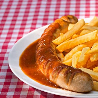 Weiße Platte mit Currywurst in Sauce, Pommes, Salatblättern, Radieschen und Besteck auf rot-weiß kariertem Tischtuch.