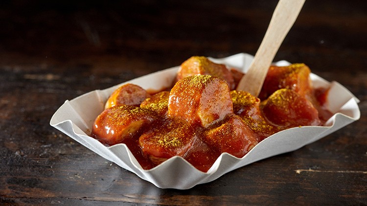 Currywurst-Stücke mit roter Sauce und gelbem Currypulver in einer weißen Papierschale, Holzspieß steckt in der Wurst.