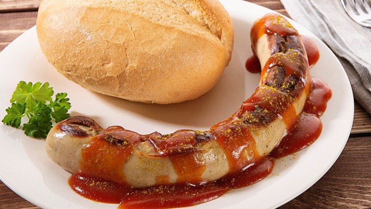 Gegrillte Bratwurst mit Currysoße und Gewürzen, serviert mit einem Brötchen und Petersilie auf einem weißen Teller.