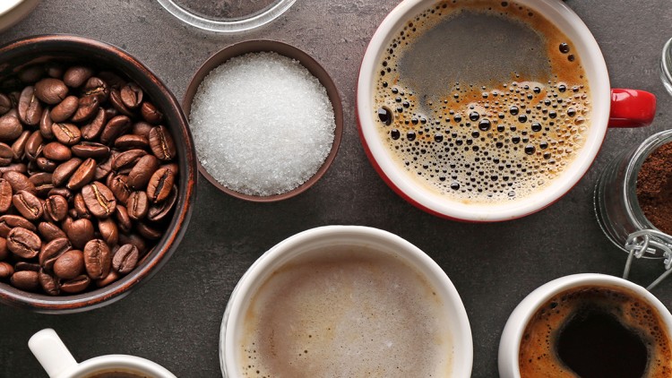Verschiedene Kaffeezutaten wie Bohnen, Zucker, Milch und Kaffeesorten in Tassen auf grauem Untergrund.