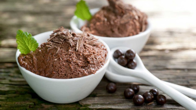 Zwei weiße Schalen mit Schokoladenmousse, garniert mit Schokoraspeln und Minzblättern, neben weißen Löffeln mit Schokokugeln.