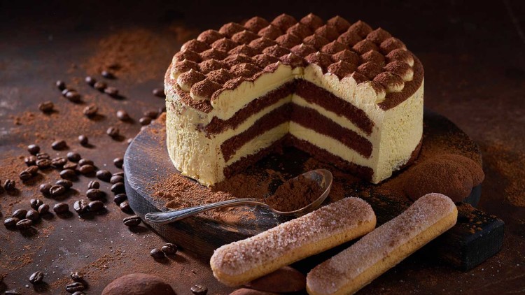 Aufgeschnittene Tiramisu-Torte mit Kakaobestäubung auf Holzbrett, daneben Löffelbiskuits, Kaffeebohnen und ein Löffel mit Kakao.