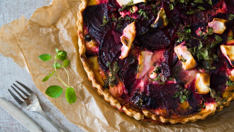 Rote-Bete-Tarte mit Ziegenkäse und Kräutern auf braunem Backpapier, daneben ein Besteck und frische grüne Kräuter.