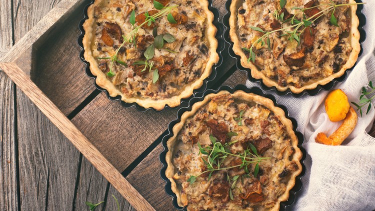 Drei Pilzquiches mit frischen Kräutern in schwarzen Backformen auf einem rustikalen Holztisch mit weißem Tuch und Pilzen.