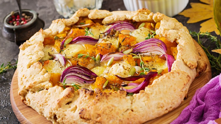 Runde Galette mit goldbraunem Rand, belegt mit Kürbisstücken, roten Zwiebeln, Käse, Kürbiskernen und frischen Kräutern.