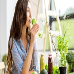 Person sitzt an einem Tisch, trinkt aus einem Glas mit grünem Inhalt und ist von frischem Gemüse und Kräutern umgeben.