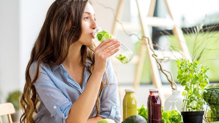Person in blau-weiß gestreiftem Hemd hält eine Glasflasche mit Wasser und Minze, Tisch mit Gemüse und Smoothies im Hintergrund.