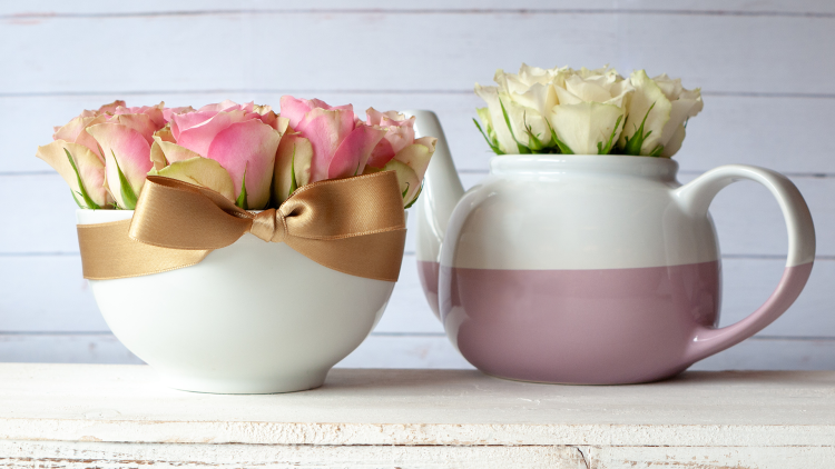 Weiße Schale mit rosa Rosen und goldener Schleife, daneben rosa-weiße Teekanne mit weißen Rosen, auf hellem Holztisch.