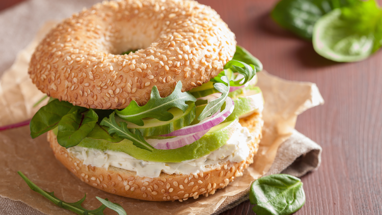 Ein Sesam-Bagel mit Frischkäse, Avocadoscheiben, roten Zwiebelringen, Rucola und Babyspinat auf braunem Papieruntergrund.