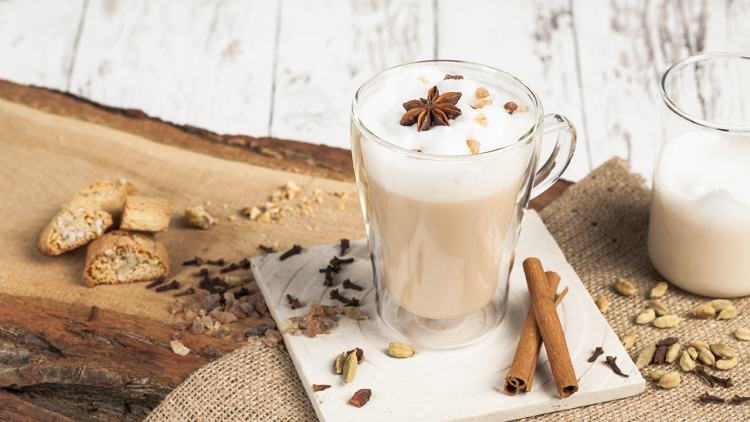 Ein Glas Milchkaffee mit Milchschaum, Sternanis und Nüssen auf einem Untersetzer, daneben Zimtstangen und Gewürze.
