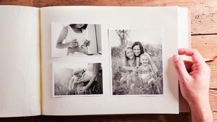 Eine Hand blättert ein Fotoalbum mit drei schwarz-weißen Fotos auf einer Holzoberfläche durch.